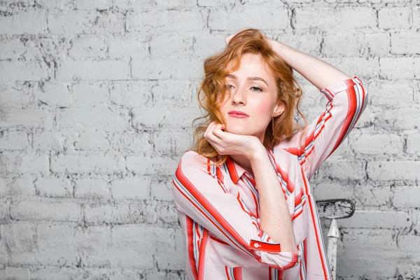 Retrato de cerca Hermosa joven estudiante con el pelo rizado rojo y pecas en su cara sentado en una pared de ladrillo en fondo gris. Vestido con una camisa de rayas rojas. Manos tocando el cabello —  Fotos de Stock