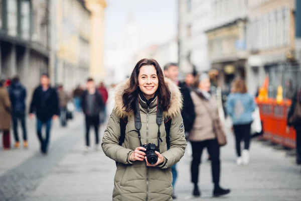 Retrato Bela jovem turista fica no fundo de uma multidão de pessoas em uma rua central em Munique, na Alemanha, no inverno. Segura uma grande câmera profissional preta e sorri — Fotografia de Stock