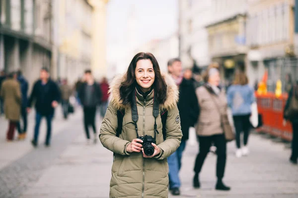 Retrato Bela jovem turista fica no fundo de uma multidão de pessoas em uma rua central em Munique, na Alemanha, no inverno. Segura uma grande câmera profissional preta e sorri — Fotografia de Stock
