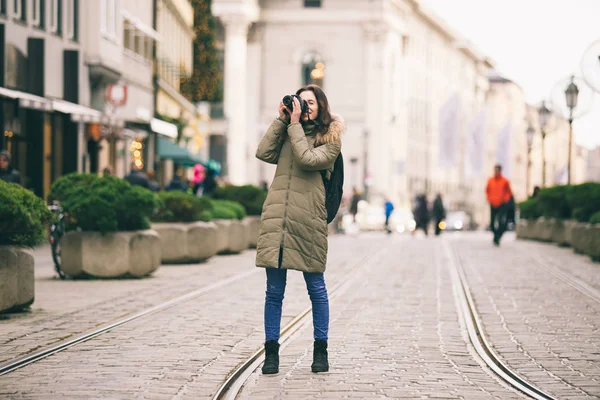 Bela jovem turista fica no fundo da rua central em Munique, na Alemanha, no inverno. Segura uma grande câmera profissional preta, tira uma foto e sorri — Fotografia de Stock