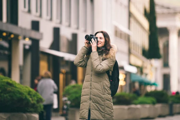 Bella giovane donna turistica si trova sullo sfondo della strada centrale di Monaco di Baviera in Germania in inverno. Tiene una grande macchina fotografica professionale nera, scatta una foto e sorride — Foto Stock