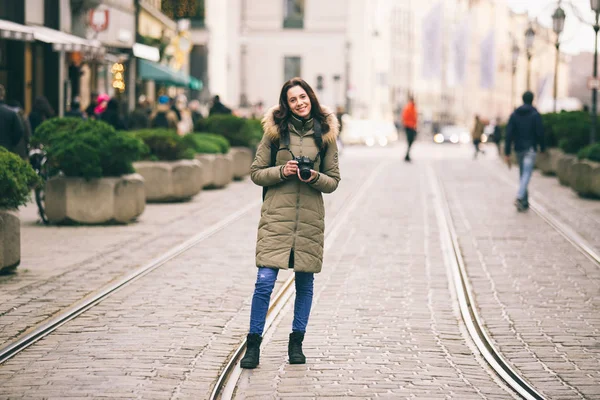 Hermosa joven turista con el pelo largo se encuentra en el fondo de la calle central de la ciudad de Munich en Alemania en invierno. Sostiene una gran cámara negra profesional, poses y sonrisas — Foto de Stock