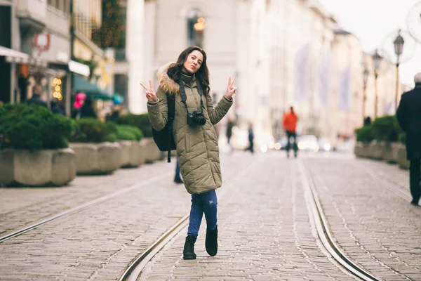 Linda jovem turista com cabelos longos fica no fundo da rua central na cidade de Munique, na Alemanha, no inverno. Mantém uma grande câmera profissional preta, poses e sorrisos — Fotografia de Stock