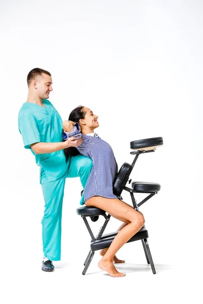 Tema masaje y oficina. joven terapeuta masculino en traje azul haciendo masaje de espalda con la rodilla apoyada en la columna vertebral de la trabajadora, mujer de negocios con camisa en la silla de masaje shiatsu para aliviar la fatiga — Foto de Stock