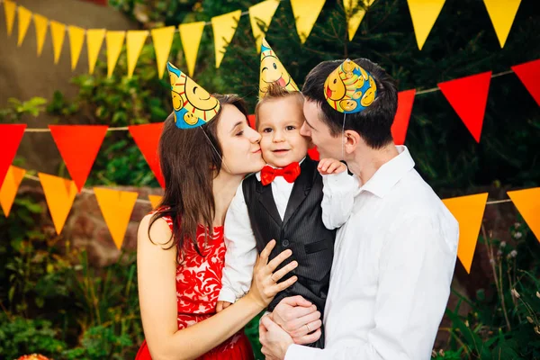 children birthday party. Family father and mother hold son of one year of birth in arms and kiss cheekin the background of park and festive decor with colored ornaments. On heads of hats, funny caps