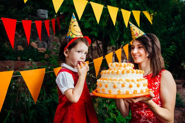 Barnens parti och tårta. Ett litet barn en rolig flicka försöker finger en tårta som mamma nästa håller i händerna. Mot bakgrund av gröna träd och festlig inredning. — Stockfoto