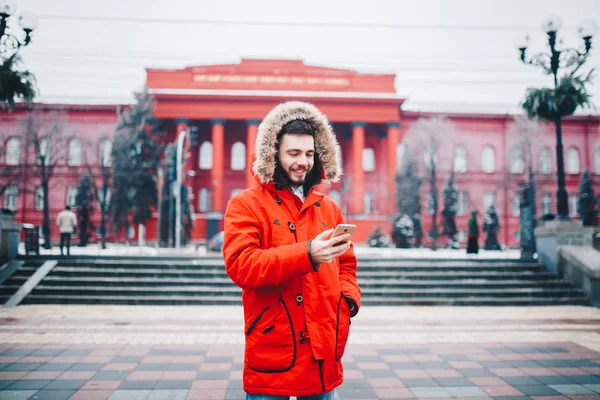 Красивый молодой парень с бородой и красной курткой в капюшоне студент пользуется мобильным телефоном, пишет, пишет корреспонденцию по телефону с улыбкой на красное здание университета или колледжа — стоковое фото