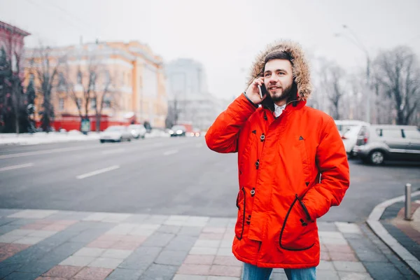 Młody facet z brodą i czerwona kurtka kaptur student wykorzystuje telefon komórkowy, trzyma w ręku w pobliżu głowy, rozmawia przez telefon z uśmiechem na tle czerwonym budynku Uniwersytetu lub college'u — Zdjęcie stockowe