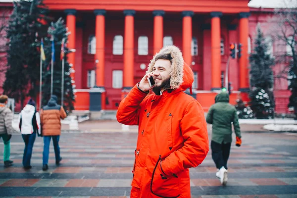 Młody facet z brodą i czerwona kurtka kaptur student wykorzystuje telefon komórkowy, trzyma w ręku w pobliżu głowy, rozmawia przez telefon z uśmiechem na tle czerwonym budynku Uniwersytetu lub college'u — Zdjęcie stockowe