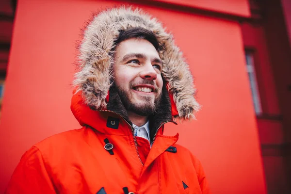 Retrato de un joven guapo, un estudiante con barba en una chaqueta roja de invierno y una capucha con piel en el fondo de la pared roja del edificio de una institución educativa —  Fotos de Stock