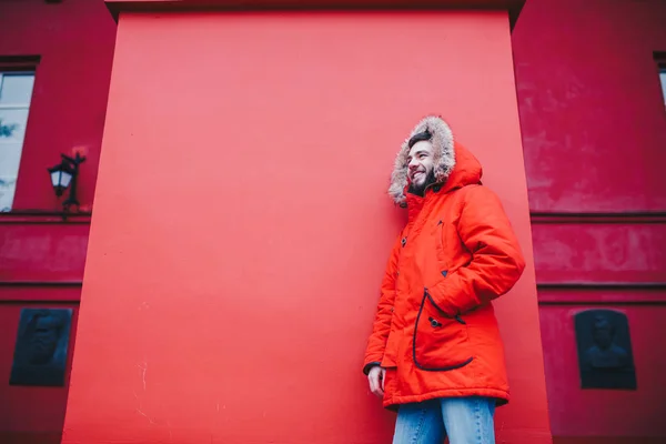 Ritratto di un giovane bel ragazzo, uno studente con la barba in giacca invernale rossa e un cappuccio con pelliccia sullo sfondo della parete rossa dell'edificio di un'istituzione educativa — Foto Stock