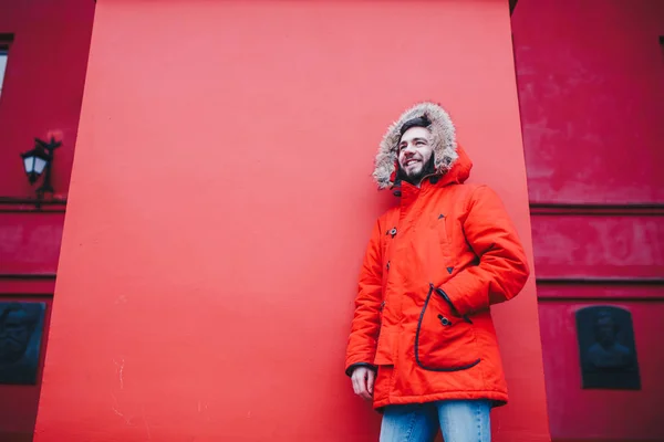 Portret młody przystojny facet, student z brodą w zimowej kurtce i kaptur z futerkiem na tle czerwone ściany budynku instytucji edukacyjnej — Zdjęcie stockowe