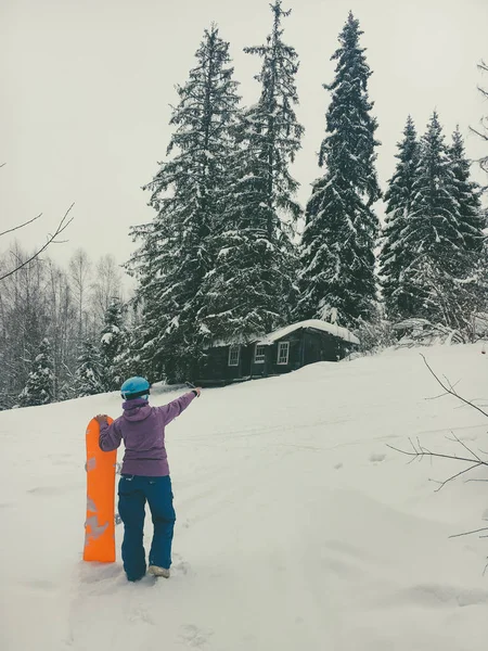 Snowboard girl dans un casque bleu se tient avec son dos tenant conseil à côté de la planche orange vif et montre la direction vers l'avant dans la forêt et regarde abandonné vieille maison en bois hutte dans la clairière — Photo