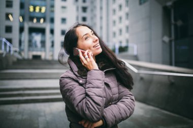genç kız cep telefonu avlu İş Merkezi konuşuyor. soğuk havalarda kış ceket giymiş uzun siyah saçlı kız arka plan binalar beton ve camdan telefonla konuşuyor