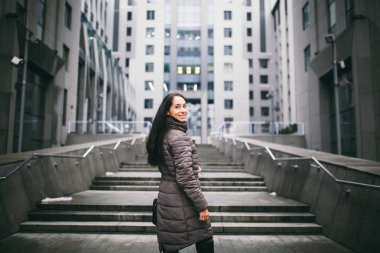 bir iş tema üzerinde bir kadın portresi. İş Merkezi arka plan, ofis binası ile cam cephe üzerinde genç beyaz esmer kız uzun ceket, palto siyah deri bir çanta içinde duruyor