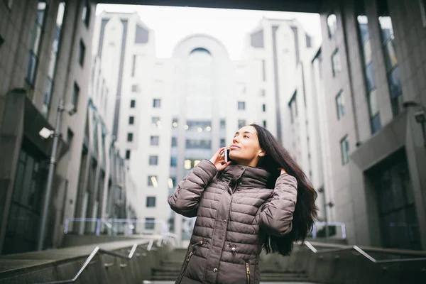 Giovane ragazza che parla al telefono cellulare nel cortile business center. ragazza con lunghi capelli scuri vestita con giacca invernale in tempo freddo parla al telefono su edifici di sfondo in vetro e cemento — Foto Stock