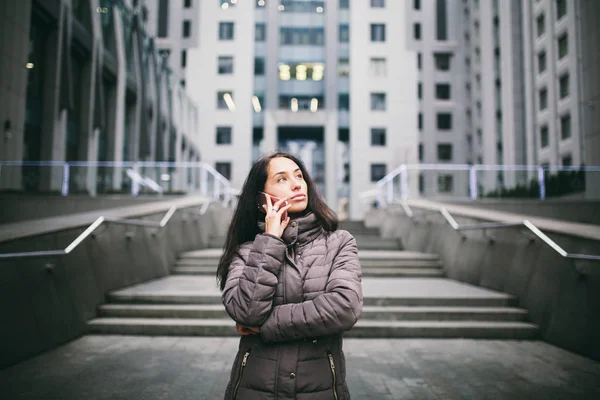 コートヤード バイ マリオット ビジネス センターで携帯電話で話している若い女の子。寒さで冬のジャケットを着た長い黒髪の少女がガラスとコンクリートで作られた背景の建物に電話で話す — ストック写真