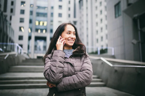 Chica joven hablando por teléfono móvil en el centro de negocios patio. chica con pelo largo y oscuro vestida con chaqueta de invierno en clima frío habla por teléfono en los edificios de fondo de vidrio y hormigón — Foto de Stock