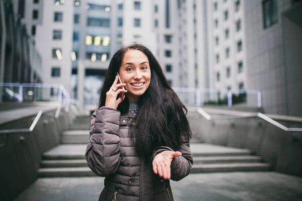 Genç kız cep telefonu avlu İş Merkezi konuşuyor. soğuk havalarda kış ceket giymiş uzun siyah saçlı kız arka plan binalar beton ve camdan telefonla konuşuyor — Stok fotoğraf