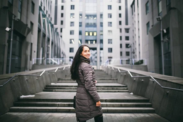 Retrato de uma mulher sobre um tema de negócios. Menina morena branca nova em jaqueta longa, casaco com saco de couro preto fica no fundo do centro de negócios, prédio de escritórios com fachada de vidro — Fotografia de Stock