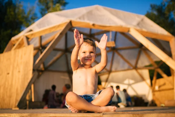 Thema Yoga und Kinder. Kaukasische Junge in blauen Shorts Kind sitzt barfuß mit den Beinen in Lotus-Pose gekreuzt und winkt mit den Armen lächelnd auf Holzboden gegen den Versammlungsort. im Sommer — Stockfoto