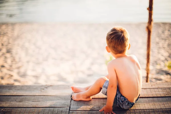 El tema es un niño y vacaciones de verano en la playa. Un niño pequeño se sienta de espaldas en un muelle de madera y admira la vista de la playa de arena y el estanque, el río. Con piernas desnudas en pantalones cortos de mezclilla azul —  Fotos de Stock