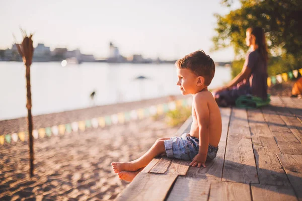 El tema es un niño y vacaciones de verano en la playa. Un niño pequeño se sienta de lado en un muelle de madera y admira la vista de la playa de arena y el estanque, el río. Con piernas desnudas en pantalones cortos de mezclilla azul —  Fotos de Stock