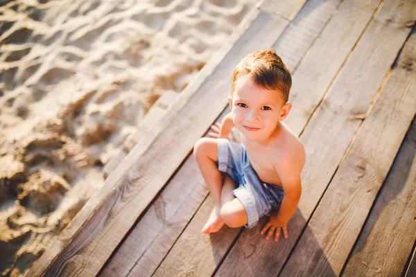 El tema es un niño y vacaciones de verano en la playa. Un pequeño niño caucásico se sienta de lado en un muelle de madera y mira a la cámara en una playa de arena y un estanque, un río. Con piernas desnudas en pantalones cortos de mezclilla azul —  Fotos de Stock