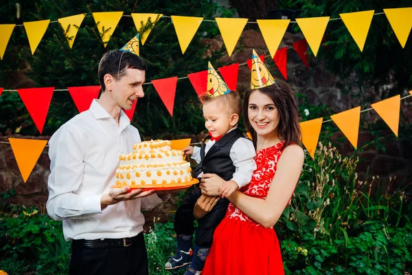 Utsätta barn födelsedagsfest, mat och godis. En ung familj firar ett år son. Pappa håller en stor tårta, Mamma håller en bebis i famnen. Baby smaker finger grädde på tårtan med tårta — Stockfoto