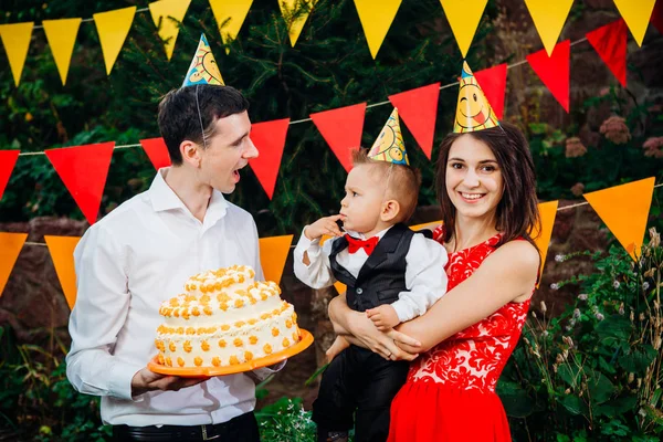 Subject children\'s birthday party, food and sweets. A young family celebrates one year of son. Dad is holding a big cake, mom is holding a baby in her arms. Baby tastes finger cream on cake with cake