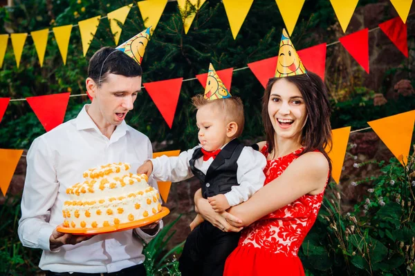 Субъект детского дня рождения, еда и сладости. Молодая семья празднует один год сына. Папа держит большой торт, мама держит ребенка на руках. Малыш пробует крем для пальцев на торте с тортом — стоковое фото