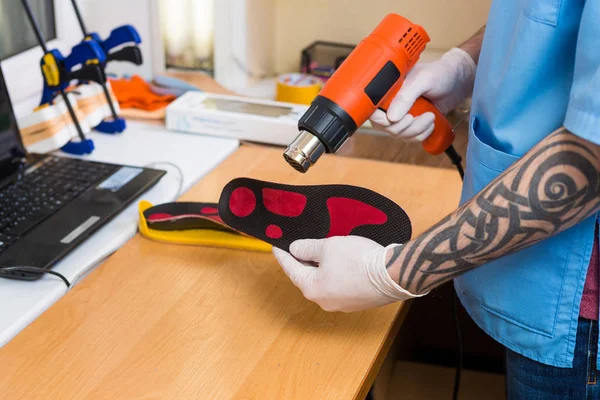 Closeup Hands of young man with tattoo in workshop dressed in blue uniform make individual orthopedic insoles. The instrument uses hairdryer to heat and deform. Theme of small business and medicine — Stock Photo, Image