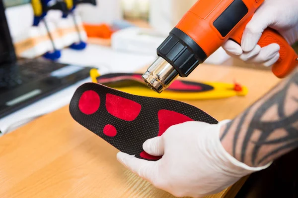 Closeup Hands of young man with tattoo in workshop dressed in blue uniform make individual orthopedic insoles. The instrument uses hairdryer to heat and deform. Theme of small business and medicine — Stock Photo, Image