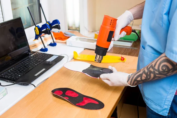 Closeup Hands of young man with tattoo in workshop dressed in blue uniform make individual orthopedic insoles. The instrument uses hairdryer to heat and deform. Theme of small business and medicine — Stock Photo, Image