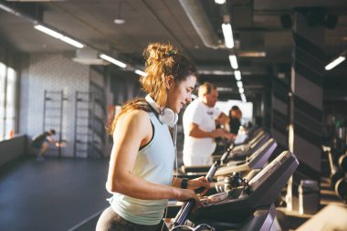 yelek ve tozluk yemyeşil kıvırcık saçlı güzel genç kadın spor salonunda koşu bandına, makine ve kilo kaybı eğitim kardiyo çalışır. Kafa büyük beyaz kulaklık üzerinde müzik dinleme. Güneşli havalarda