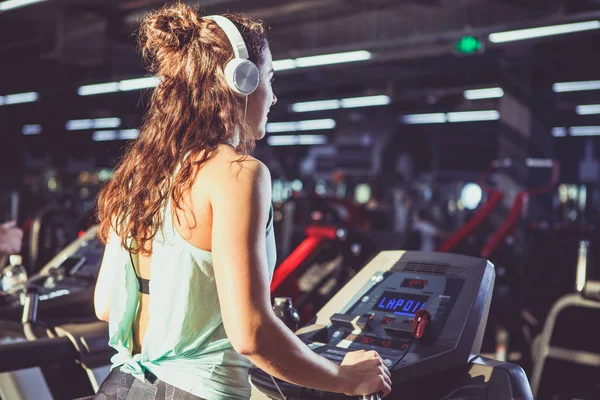 Back view. beautiful woman with lush curly hair in vest and elk runs in gym on treadmill, cardio training machine and weight loss. On head big white headphones, listening music. Sunny weather