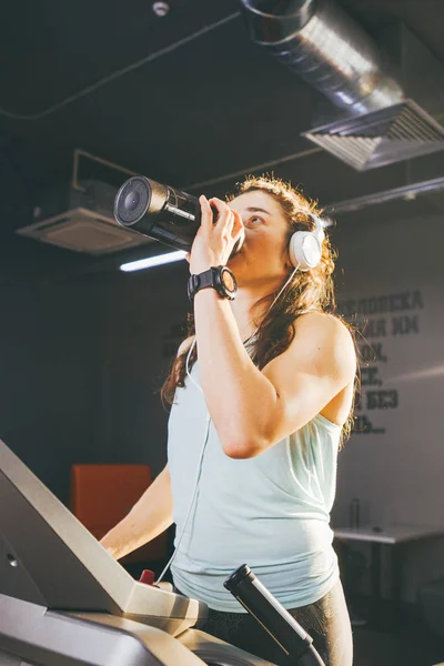 Sports nutrition and exercise. A beautiful young woman drinks protein from a bottle, a black shaker in the gym running on a treadmill, at the hands of a sports watch, in sunny weather