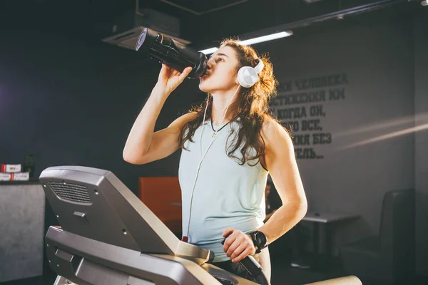 Nutrición deportiva y ejercicio. Una hermosa joven bebe proteína de una botella, una coctelera negra en el gimnasio corriendo en una cinta de correr, a manos de un reloj deportivo, en un clima soleado — Foto de Stock
