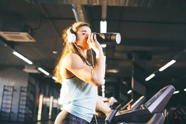Sports nutrition and exercise. A beautiful young woman drinks protein from a bottle, a black shaker in the gym running on a treadmill, at the hands of a sports watch, in sunny weather