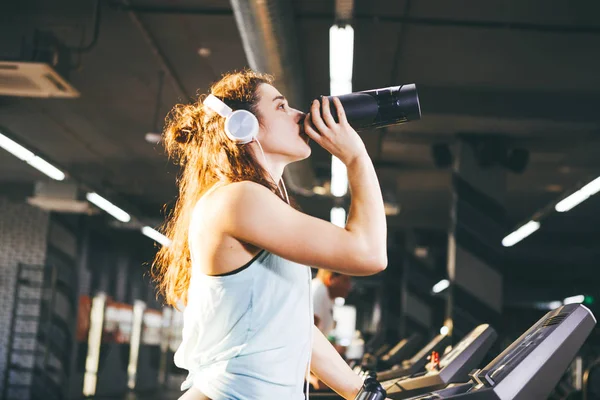 Sports nutrition and exercise. A beautiful young woman drinks protein from a bottle, a black shaker in the gym running on a treadmill, at the hands of a sports watch, in sunny weather