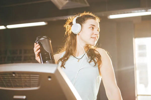 Nutrición deportiva y ejercicio. hermosa mujer tiene proteína en la mano en botellas, agitador, escucha música en auriculares en la cabeza en el gimnasio corriendo en la cinta de correr, a manos de reloj deportivo, en tiempo soleado — Foto de Stock