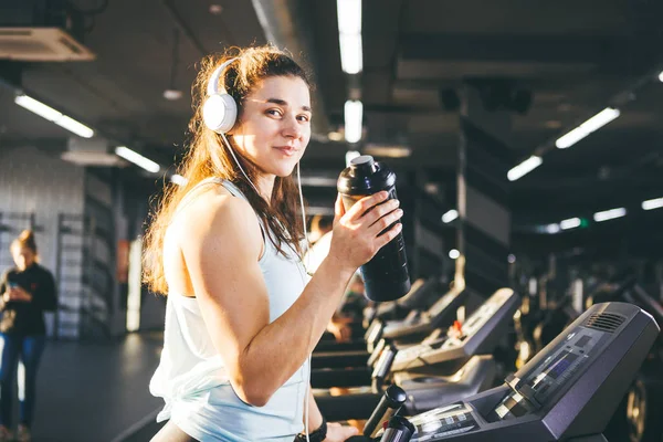 Nutrición deportiva y ejercicio. hermosa mujer tiene proteína en la mano en botellas, agitador, escucha música en auriculares en la cabeza en el gimnasio corriendo en la cinta de correr, a manos de reloj deportivo, en tiempo soleado — Foto de Stock