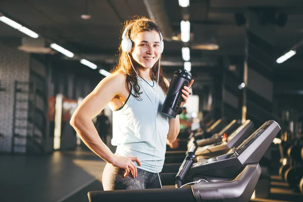 Sports nutrition and exercise. beautiful woman holds protein in her hand in bottles, shaker, listens music in headphones on head in gym running on treadmill, at hands of sports watch,in sunny weather