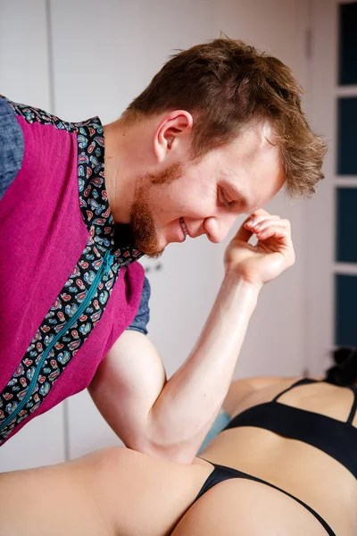 Um terapeuta de massagem masculino com barba em um terno vermelho faz uma massagem anti-celulite para uma menina em roupa interior preta com a ajuda de um cotovelo no salão de beleza em uma mesa de massagem. — Fotografia de Stock