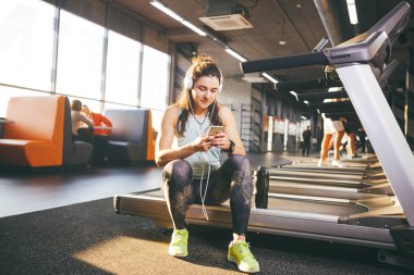 Oturmuş, güneşli havalarda spor salonu zemin karşı koşu bandı üzerinde eğitim sonra dinlenme güzel genç beyaz kız sporcumuz. Hasta beyaz kulaklık, müzik dinler elinde tutan telefon