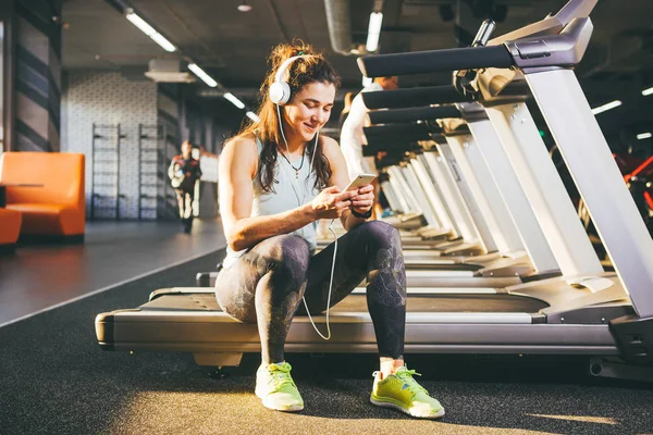 Hermosa joven chica caucásica deportista sentado, descansando después de entrenar en la cinta de correr contra el telón de fondo del gimnasio en el clima soleado. Escucha música en auriculares blancos enfermos, en la mano sostiene el teléfono — Foto de Stock