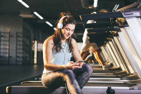 Hermosa joven chica caucásica deportista sentado, descansando después de entrenar en la cinta de correr contra el telón de fondo del gimnasio en el clima soleado. Escucha música en auriculares blancos enfermos, en la mano sostiene el teléfono — Foto de Stock