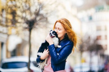Genç redhaired beyaz bir kadın çiller ile tutar ve öper, siyah ve beyaz kucaklayan tüylü köpek Chihuahua cins. mavi deri ceket giymiş kız Avrupa'da baharda sokakta duruyor