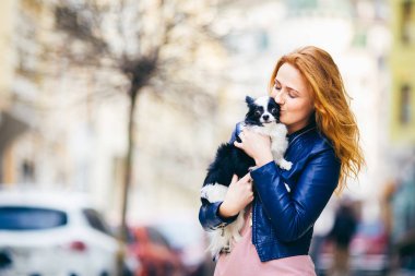 Genç redhaired beyaz bir kadın çiller ile tutar ve öper, siyah ve beyaz kucaklayan tüylü köpek Chihuahua cins. mavi deri ceket giymiş kız Avrupa'da baharda sokakta duruyor