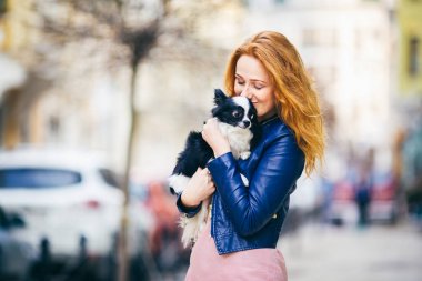 Genç redhaired beyaz bir kadın çiller ile tutar ve öper, siyah ve beyaz kucaklayan tüylü köpek Chihuahua cins. mavi deri ceket giymiş kız Avrupa'da baharda sokakta duruyor